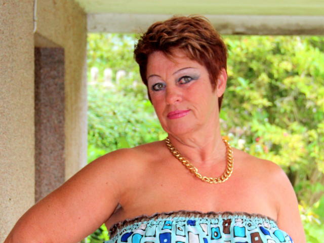 Bettina, com cabelo castanho curto, maquiagem azul nos olhos, batom rosa, colar de corrente dourada, blusa sem alças com padrão azul e marrom, em pé do lado de fora, olhando ligeiramente para a direita..