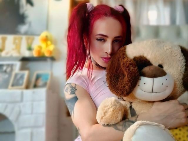 YeiSela, com maria-chiquinhas vermelhas, segurando um cachorro de pelúcia marrom e branco, vestindo uma blusa rosa, mostrando tatuagens em seu braço, sorrindo para a câmera. Seus seios estão visíveis..