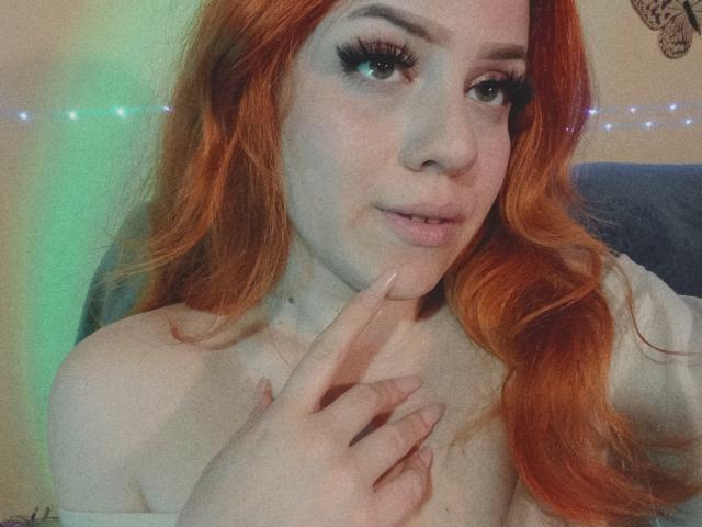 SpicyGrace, con i capelli rossi, indossa lunghe ciglia, tocca il mento con un dito, mostrando le spalle scoperte, e sorride dolcemente. Ha un viso carino e sensuale..