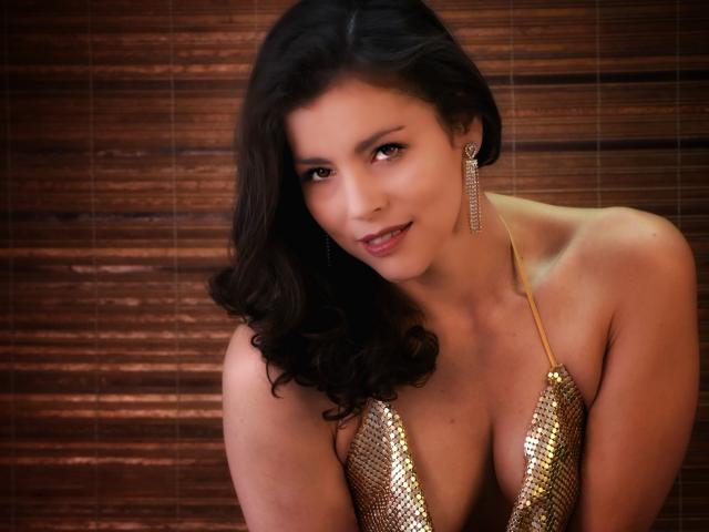 Scottie with wavy dark hair, gold sequined top, showing her tits, wearing long dangling earrings, smiling softly, brown wooden background.