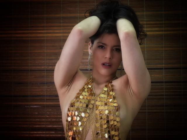 Scottie with hands in dark hair, wearing gold sequined halter top, showing tits, looking at camera with slightly open mouth, wooden background.