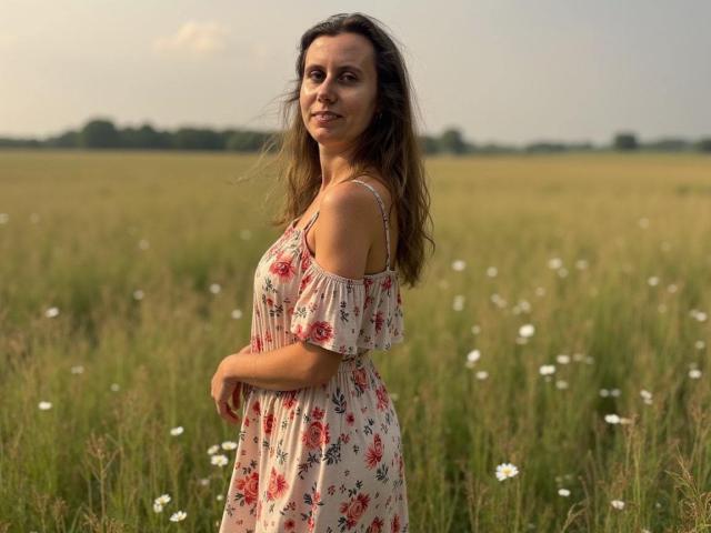 LizzySparkle, with small tits, wearing a floral dress, stands in a grassy field, her brown hair blowing, hands on her belly, smiling, white flowers in the background.