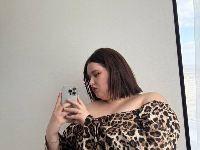Jessica Joye, avec des cheveux bruns mi-longs, portant un haut léopard à épaules dénudées, prend un selfie devant un miroir avec un téléphone vert, le regard baissé et des lèvres rouges..