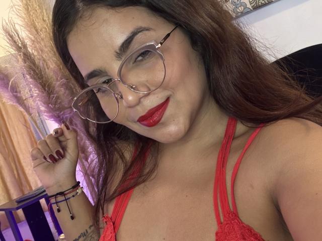 SexxyNikole with red lipstick, glasses, and red lingerie, showing her tits, smiling, with long brown hair and maroon nails, holding her hair. Her wrist has bracelets.