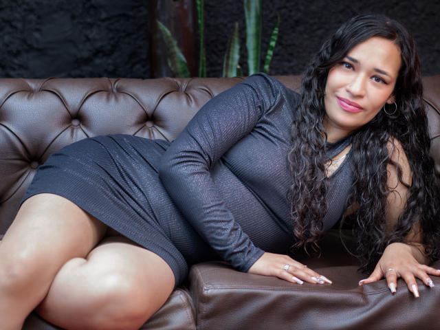 ZoeyMoretti with curly black hair, wearing a shiny gray dress, laying on a brown leather couch, showing her ass, smiling with pink lipstick, and gold hoop earrings.