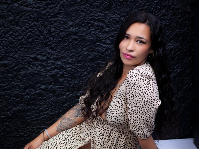 ZoeyMoretti with long black wavy hair, pink lipstick, tattoo on right arm, wearing a beige leopard-print dress, standing against a textured black wall, looking at camera.