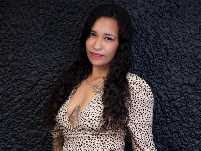 Zoey Moretti, avec ses longs cheveux noirs bouclés, sa peau brune et son rouge à lèvres rose, porte un haut à imprimé léopard à décolleté plongeant, qui révèle ses seins et son tatouage sous sa poitrine. Elle sourit légèrement..