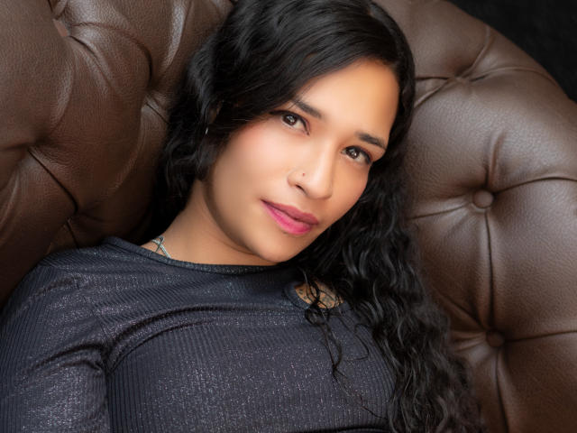 Zoey Moretti, con cabello negro rizado, ojos marrones y lápiz labial rosa, vestida con una blusa negra brillante, sentada en una silla de cuero marrón con capitonado, mirando directamente a la cámara..