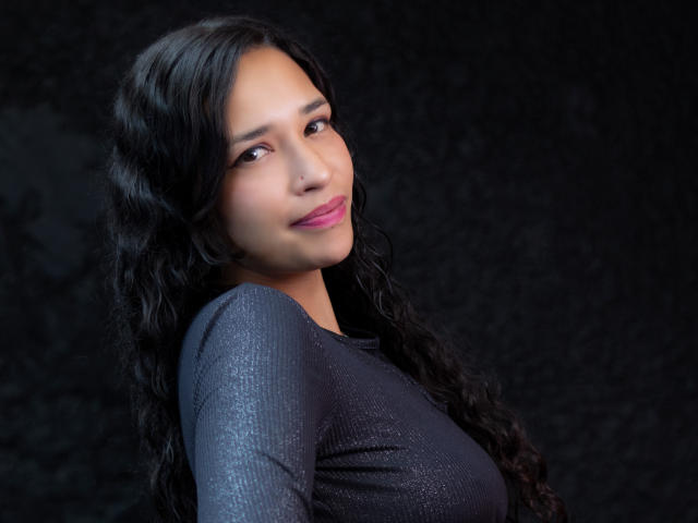 Zoey Moretti, com longos cabelos negros ondulados, vestindo uma blusa brilhante, de cor cinza escura, sorrindo discretamente, com batom rosa e uma expressão suave e confiante. Seus seios não estão visíveis..