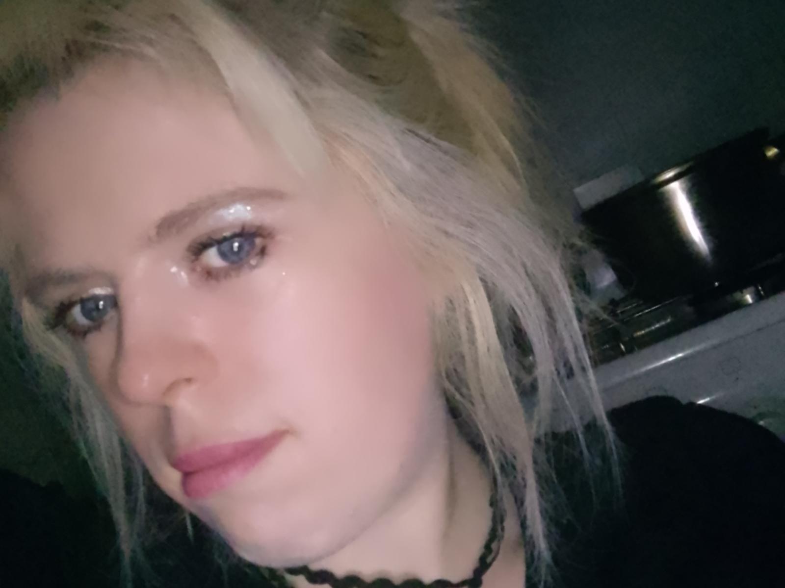 CharleSugar has pale skin, blue eyes, and blonde messy hair. She's wearing black lace and has a slight pink lip. Her face is shiny, with a subtle smile.