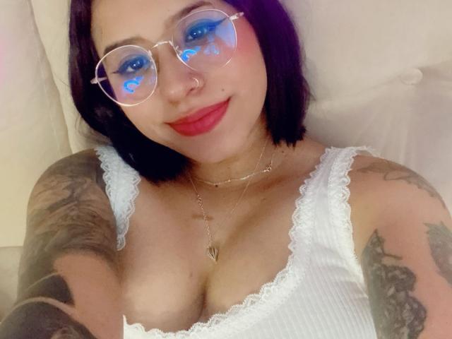 AshleyMayer with blue eye makeup, clear glasses, red lipstick, white lace top, tattoos on arms, and a heart necklace, smiling at the camera. Boobs visible, short black hair.