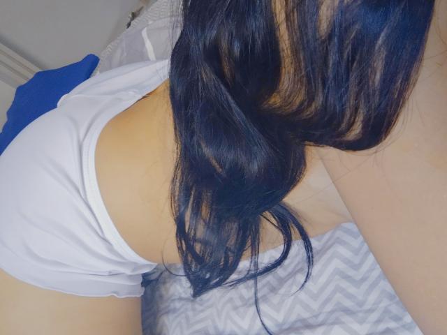 MorenaSexual with long black hair, white shirt, and blue cloth, on bed, showing her back and butt. Chevrons on bed. Soft lighting.