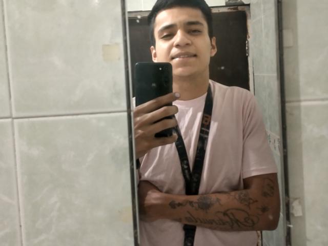 AngeloHott69 with short black hair, smiling, wearing a pink shirt and black lanyard, holding a phone, tattooed arms, mirror selfie, medium skin tone, casual.