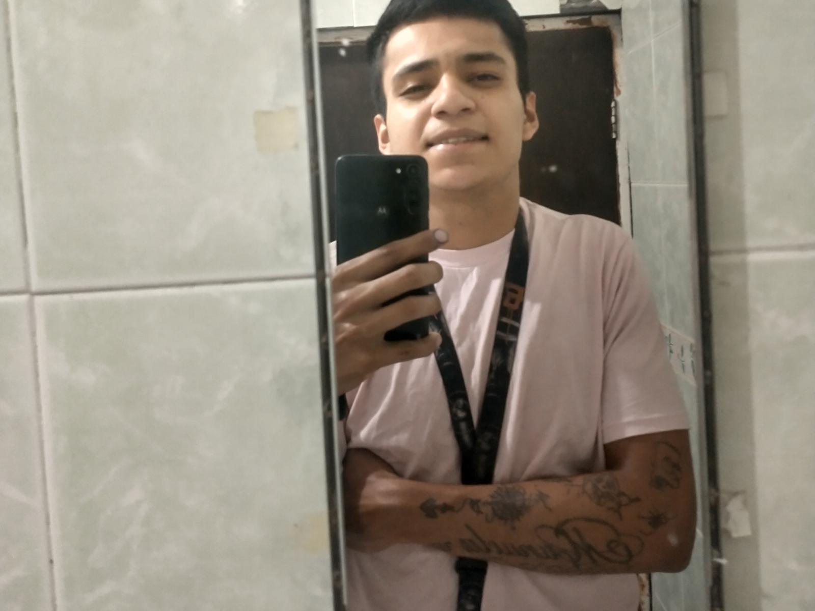 AngeloHott69 with short black hair, smiling, wearing a pink shirt and black lanyard, holding a phone, tattooed arms, mirror selfie, medium skin tone, casual.