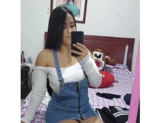 AlexaJackson69 with long black hair, white off-shoulder top, and denim overalls, taking a mirror selfie, showing her bare shoulders and small tits, holding a phone in a bedroom.