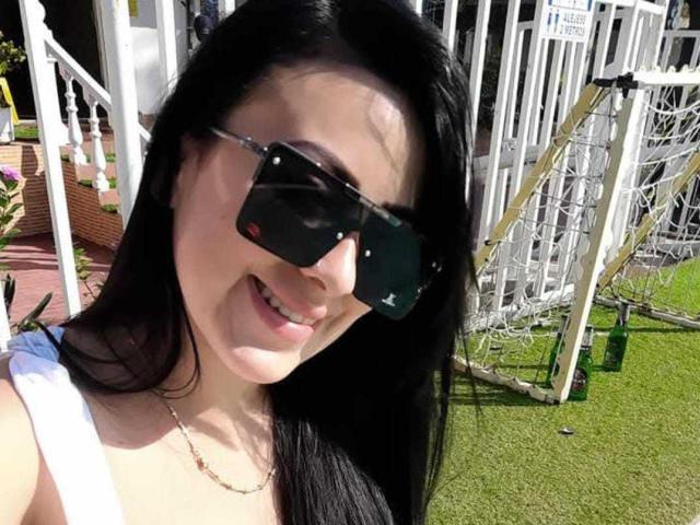AlexaJackson69 with long black hair, smiling, wearing dark sunglasses and a white top, showing her cute face and slightly visible ass in a sunny outdoor selfie.