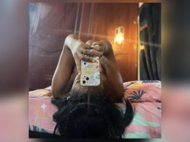 TiffanySammy, dark-skinned, with braided hair, taking a selfie, lying on a bed with colorful, patterned sheets, holding a phone with "Dad" sticker, in a dimly lit room.