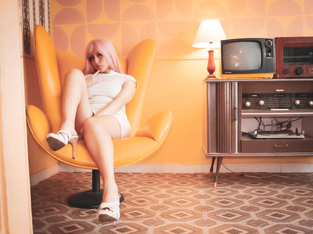 CatalinaRogers with pink hair, white dress, and high heels, sitting in an orange chair, legs crossed, showing her white-heeled toes and a hint of her white panties.