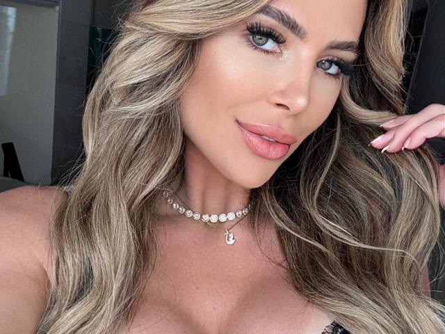 NicolyGF has blonde wavy hair, blue eyes, and pink lipstick. She's wearing a diamond necklace and has her hand in her hair, showing off long, manicured nails.