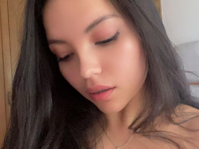 IssaPuasini's face, looking down with eyes closed, has long black hair, pink eyeshadow, and full pink lips. She's wearing a thin gold necklace, with a soft, natural skin tone.
