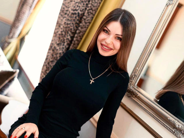 BerlinerGirl89-hot with long brown hair, smiling, wearing a black turtleneck, silver cross necklace, and sitting near a mirror, showing her tits subtly through the tight top.