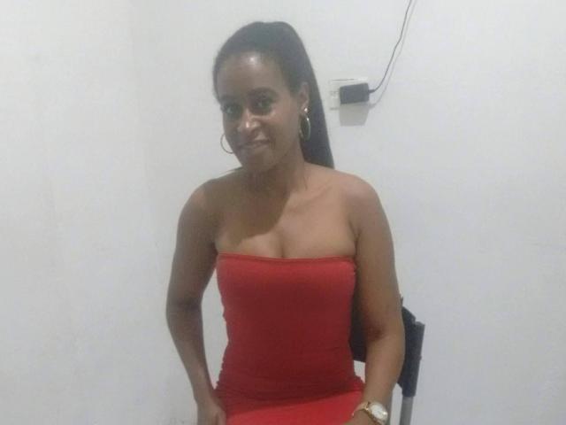 LilaLoveHot, con la pelle scura, indossa un abito rosso senza spalline che mette in mostra il seno, orecchini a cerchio dorati e un orologio dorato, sorride leggermente e sta in piedi contro un muro bianco..