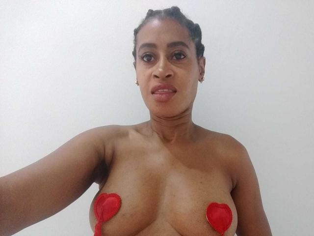 LilaLoveHot with dark brown skin, braided hair, small hoop earrings, and red heart pasties covering her tits, standing against a plain white background.