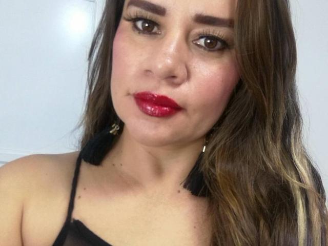 CharllotI has glossy red lips, thick brown eyebrows, long brown hair, and wears black tassel earrings with a black halter top. Her brown eyes are slightly upturned.