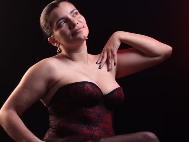BulmaHot with medium tits, wearing a red lace strapless dress, has dark hair pulled back, right arm raised, black background, and a confident, slightly smirking expression.