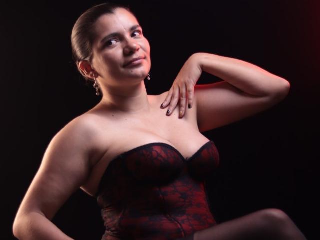 BulmaHot with dark red lace corset, showing off her tits, arms raised, dark nails, smirking, black background, red light on right, ear cuff, hair pulled back.