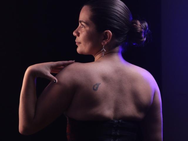 BulmaHot with her back to the camera, dark hair in a bun, strapless black dress, small tattoo on left shoulder, earrings, right hand on shoulder, purple-blue lighting.