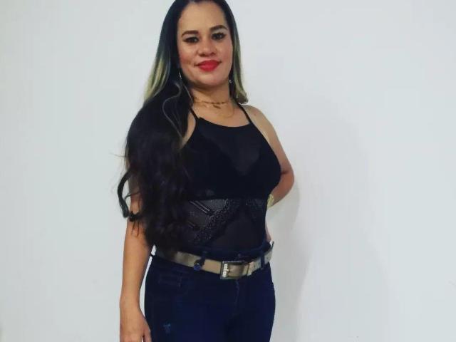 CharllotI with long black and green hair, red lipstick, black lace tank top, blue jeans, and a beige belt, standing and smiling. Boobs are medium-sized and visible.