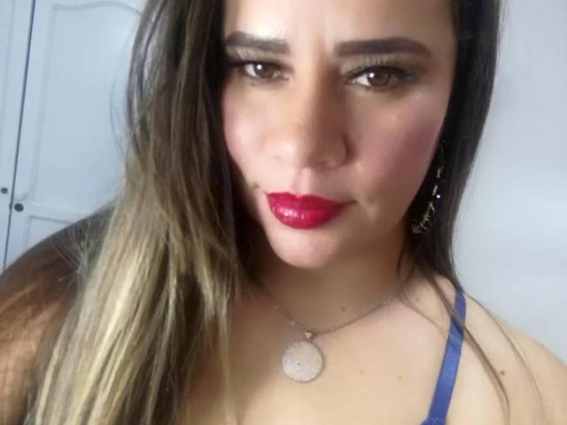 CharllotI with red lipstick, brown eyes, and long brown hair, wearing a blue top and gold earrings, looking directly at the camera with a slight smile.