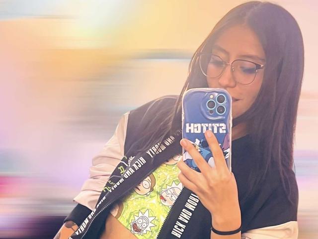 EviieGothel with long black hair, glasses, and a black/white jacket, holding a blue phone with "HOTTE" case, taking a selfie, showing a yellow green bra with cartoon.