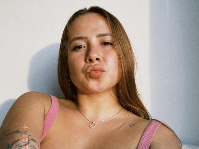 NatSorian has pouty lips, light brown hair, pink tank top, heart necklace, tattoos on shoulder, and a cute face. She's looking at the camera with a relaxed expression.