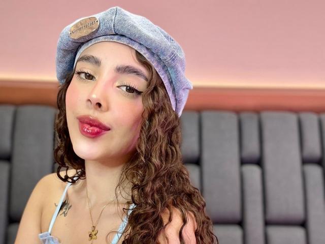 TianaVera with curly brown hair, wearing a blue beret, red lipstick, and a light blue top, showing off her tits with a subtle smile, sitting in a dark booth.