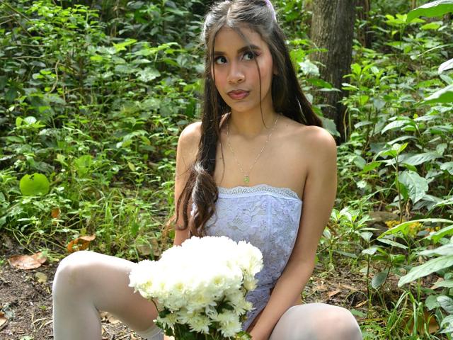 Adaallen mit langen, braunen Haaren, die ein trägerloses Spitzenoberteil und weiße Strumpfhosen trägt, hält einen weißen Blumenstrauß und sitzt in einem Wald. Sie schaut mit einem leichten Lächeln in die Kamera..