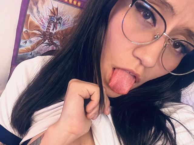 EviieGothel with black hair, glasses, and nose ring, sticking out her pink tongue, pulling up her white shirt to reveal her small tits. Tattoo on right forearm.