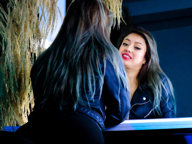 AntonellaRiquel with long, wavy blonde hair, black leather jacket, and red lipstick, leaning forward, looking in mirror, smiling, showing her ass, reflection visible.