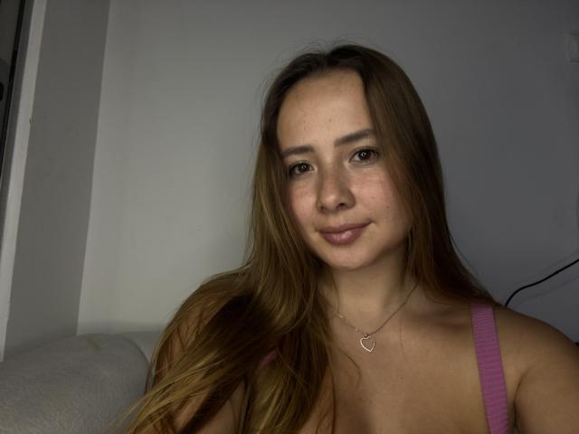 NatSorian met lang bruin haar, een roze top, een kleine hartketting, een lichte glimlach en natuurlijke borsten, in gedimd licht, met een schattig en ontspannen gezicht..