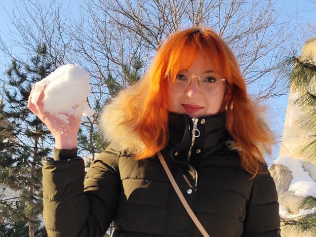 MoonBlossom with bright orange hair, wearing a black puffy coat with fur trim, holding a snowball in her right hand, smiling with glasses on, in a snowy outdoor setting.