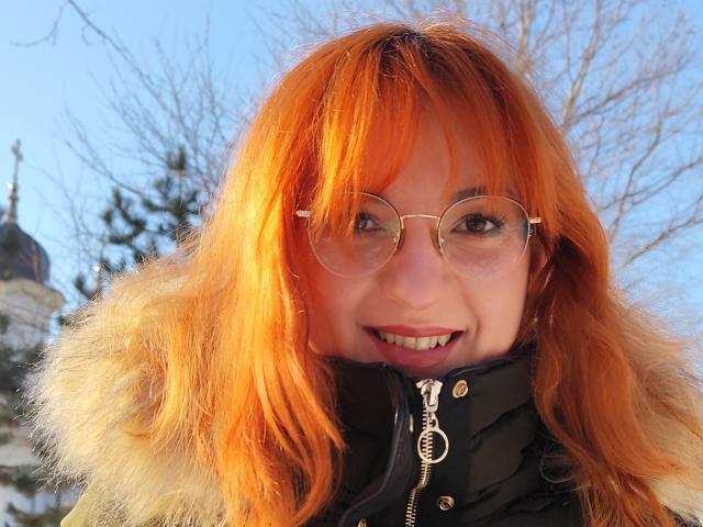 MoonBlossom has bright orange hair, round glasses, and a cute smile, wearing a puffy black jacket with a fur collar, in a sunny, wintery outdoor setting.