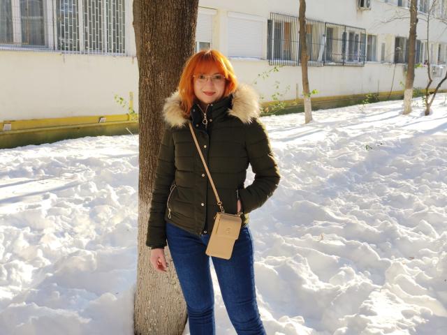 MoonBlossom with orange hair, green puffy jacket, fur collar, blue jeans, tan crossbody bag, standing in snow, leaning on tree, smiling, sunlight, winter, hands in pockets.