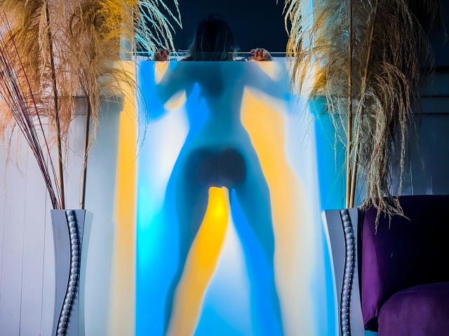 AliceHoe stands naked, back to camera, blue and yellow light highlighting her ass, silhouette against translucent curtain, flanked by potted reeds, purple chair to right.