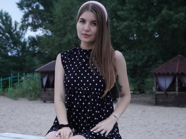 EllenMays, with long brown hair, wearing a black polka dot dress, sitting on a sandy beach, has a small tattoo on her right arm, looking slightly shy.