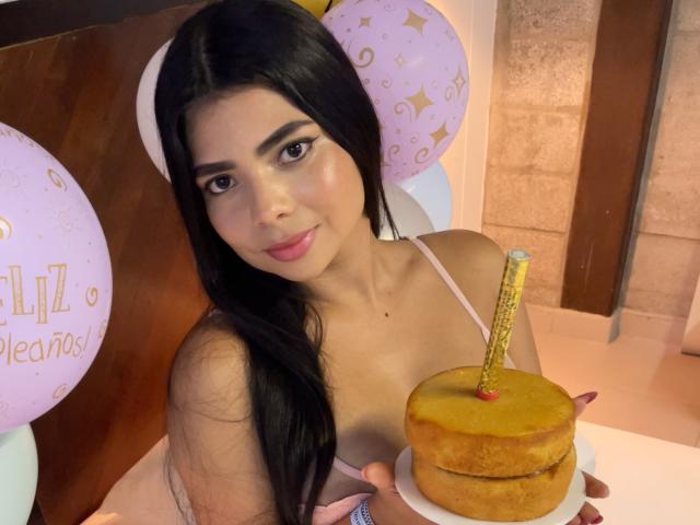 BellaClaire with long black hair, pink lingerie, holding a yellow cake with a gold candle, smiling at the camera, balloons in the background.