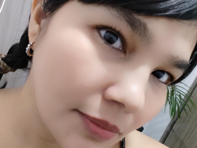 PrittyAlicia's face with light skin, black hair in a braid, brown eyes, and subtle makeup, showing a gentle smile with pink lips. She has small earrings and a slightly flushed complexion.