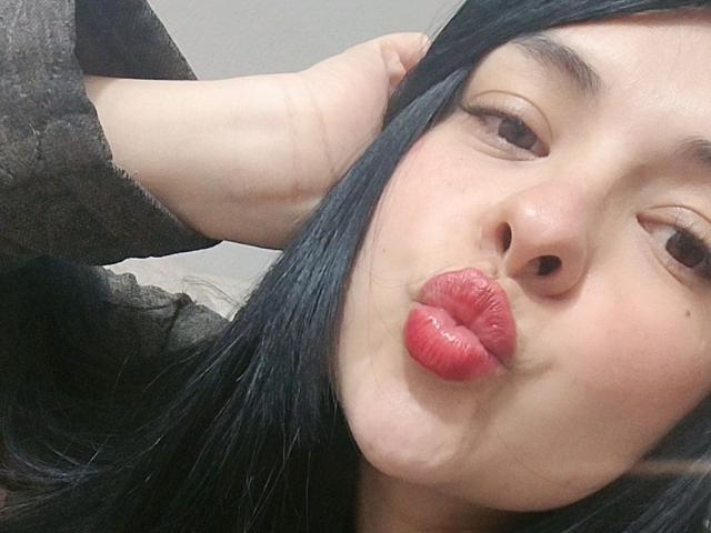 JulyMia's pouty red lips are puckered up, making a kissy face. She's got black hair, a hand on her head, and is wearing a dark gray sweater.