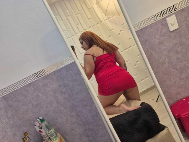 KatiaRodriguez with red dress, kneeling, showing her ass in mirror. She has long brown hair, light skin, and is in a purple-walled room with a white door.
