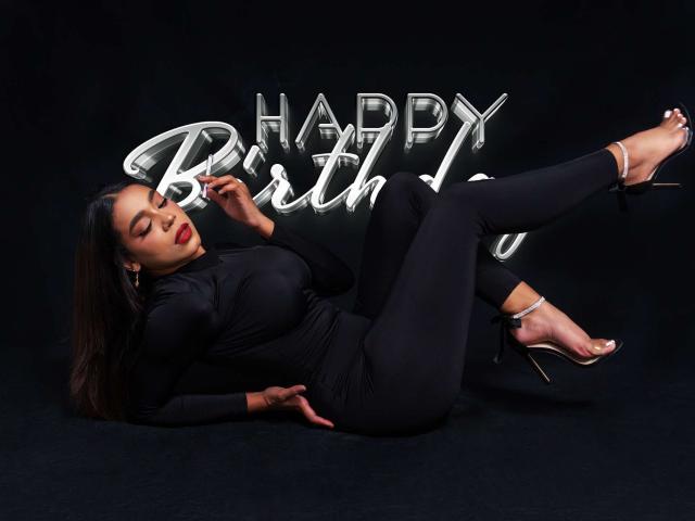 SamanthaStele with big tits, lying on black floor, wearing tight black dress, high heels, and silver earrings, legs up, looking at camera, "Happy Birthday" neon sign in background.